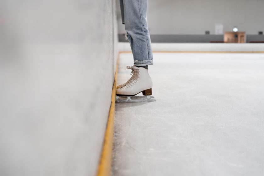 Eine Person steht mit Schlittschuhen auf einer Eisbahn. Es sind nur die Beine und die Schuhe der Person zu erkennen.