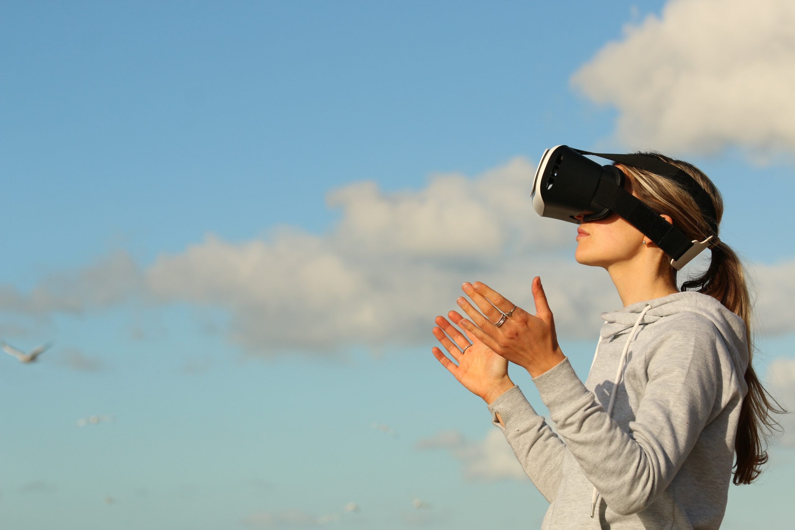 Eine Frau mit VR-Brille blickt in Richtung eines blauen Himmels. Sie hält beide Arme gebetsartig nach oben.