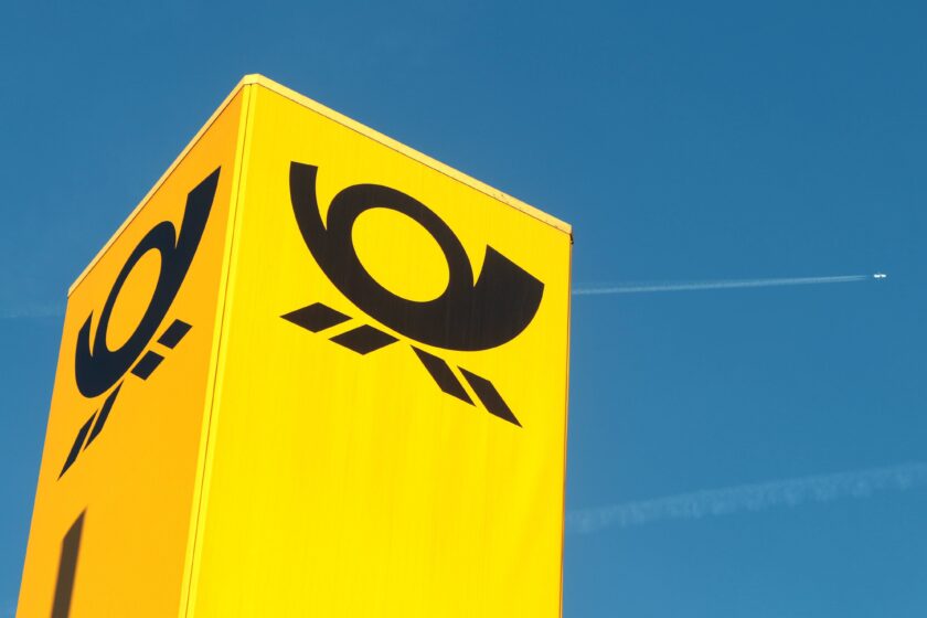 Gelber Briefkasten der Deutschen Post vor blauem Himmel mit Flugzeug am Himmel
