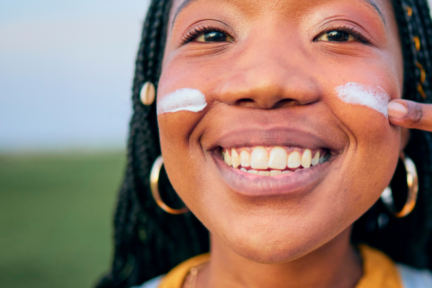eine lächelnde schwarze junge Frau malt sich einen weißen Streifen Sonnencreme auf die linke Wange, auf der rechten Wange hat sie bereits einen Streifen Sonnencreme