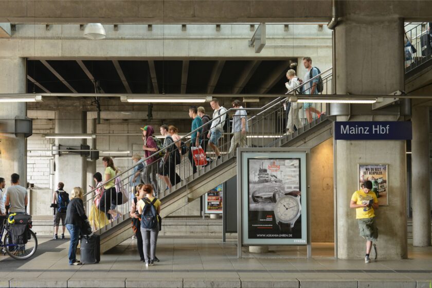 Mehrere Reisende laufen eine Treppe am Mainzer Hauptbahnhof hinunter, einige Menschen stehen und warten bereits am Bahnsteig auf ihren Zug