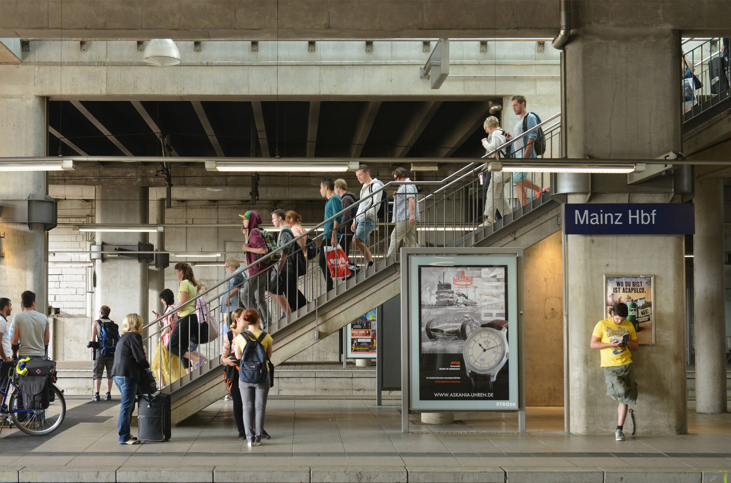 Mehrere Reisende laufen eine Treppe am Mainzer Hauptbahnhof hinunter, einige Menschen stehen und warten bereits am Bahnsteig auf ihren Zug