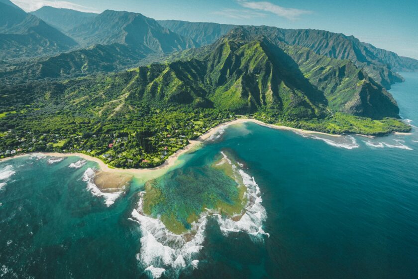 Eine Luftaufnahme von einer hawaiianischen Küste: man sieht grün bewachsene Berge, die bis ans Meer heranreichen, Berge im Hintergrund, einen schmalen Küstensandstreifen und blaues Meer, das sich schaumig an der Küste bricht