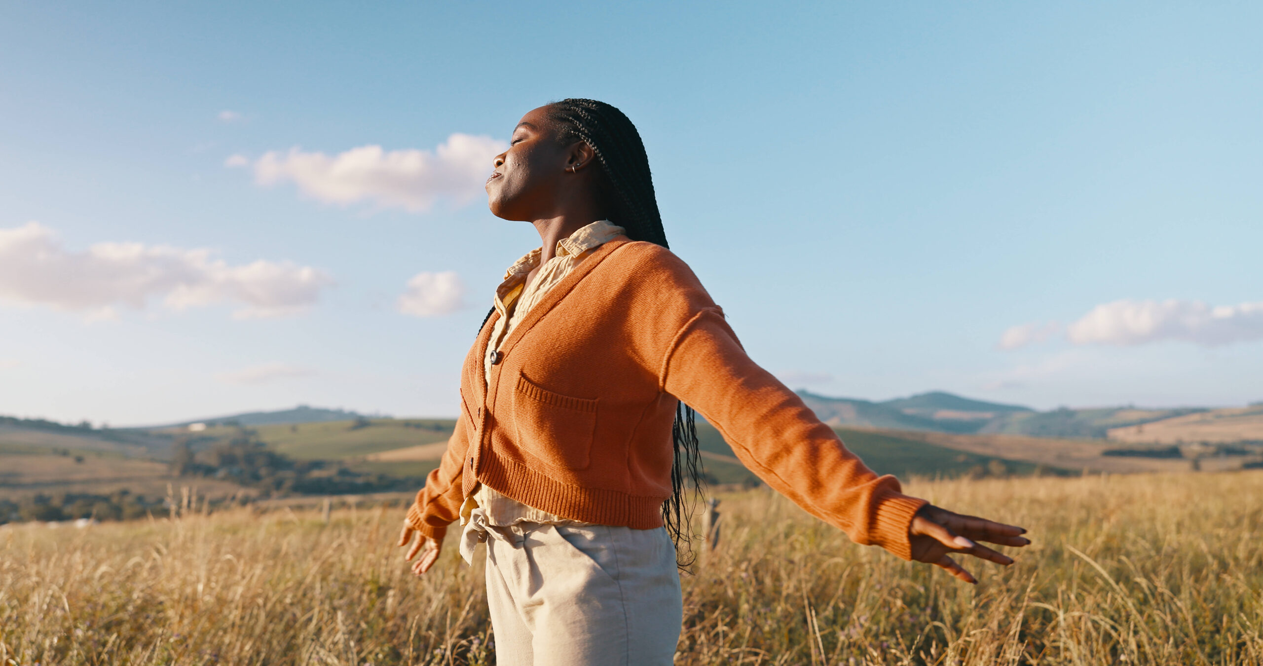 eine glückliche Schwarze Frau in einem orangefarbenen Cardigan steht mit ausgebreiteten Armen in einem Kornfeld, im Hintergrund ein blauer Himmel und ein paar kleine Wolken