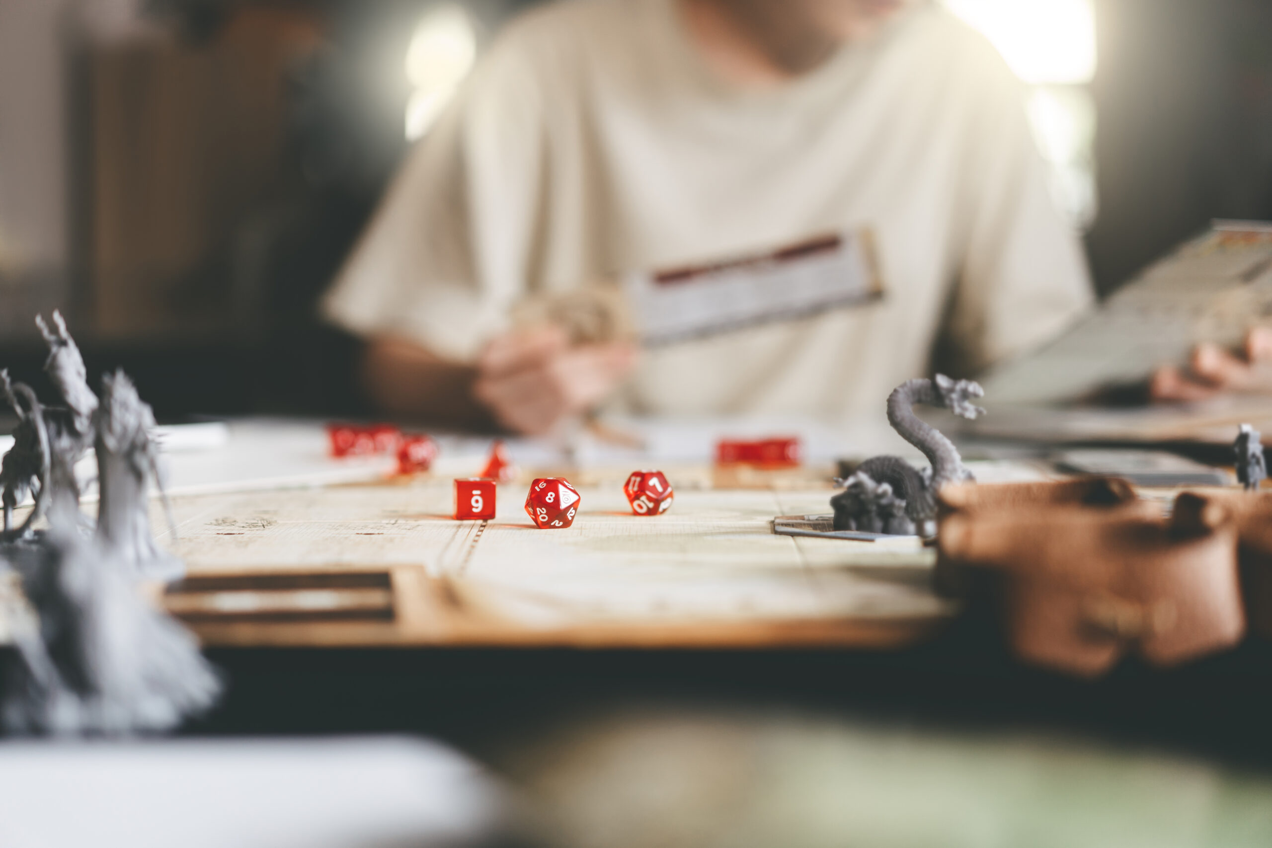 im Vordergrund: ein Tisch, auf dem eine Drachenminiatur und ein Set D&D-Würfel zu sehen ist. Im Hintergrund: ein Mensch in weißem T-Shirt, der einen Charakterbogen in der Hand hält und etwas nachliest