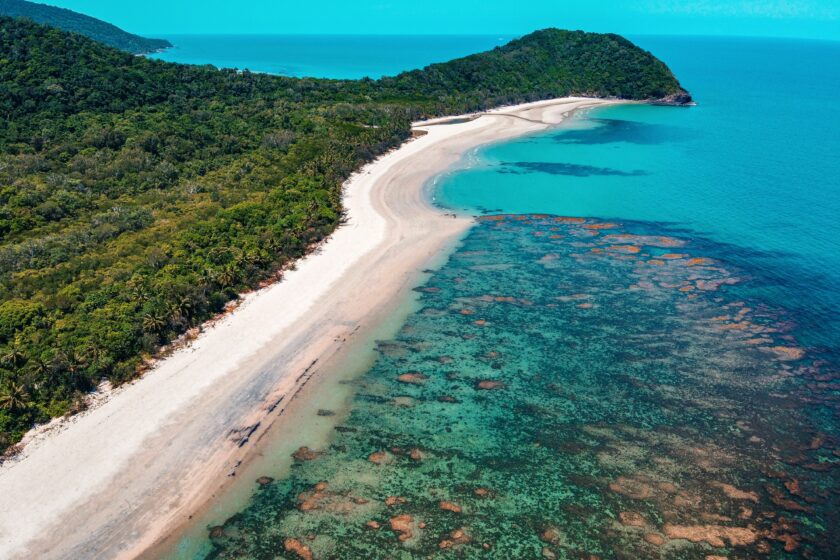 Eine Luftaufnahme, auf der man zur linken Seite den Daintree Regenwald in Australien am Cape Tribulation sieht und auf der rechten Seite das Meer und ein großes Korallenriff