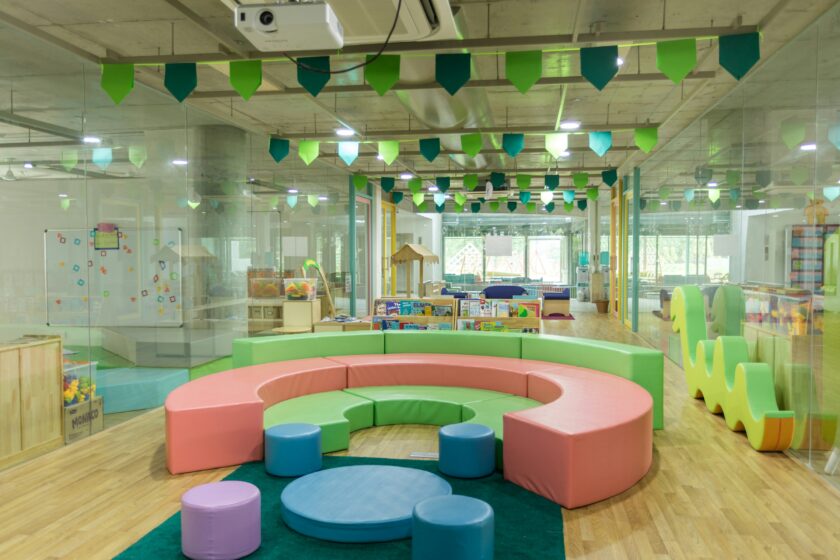 Innenansicht eines leeren Kindergartens mit bunten kreisrunden modularen Sofas, von der Decke hängenden grünen Wimpeln und Holzspielzeug und Büchern im Hintergrund