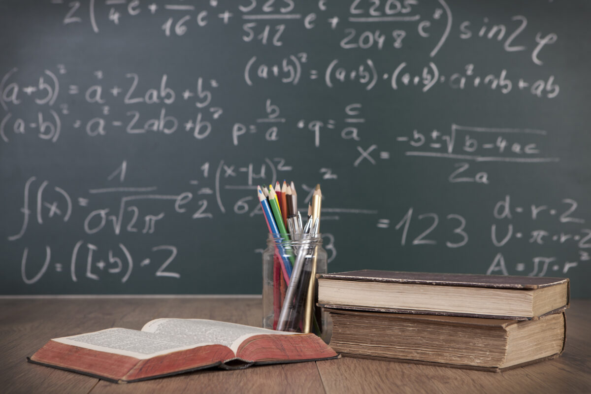 Im Vordergrund: Bücher und ein Glas voller Stifte auf einem Tisch; im Hintergrund: eine Tafel beschriftet mit mathematischen Gleichung