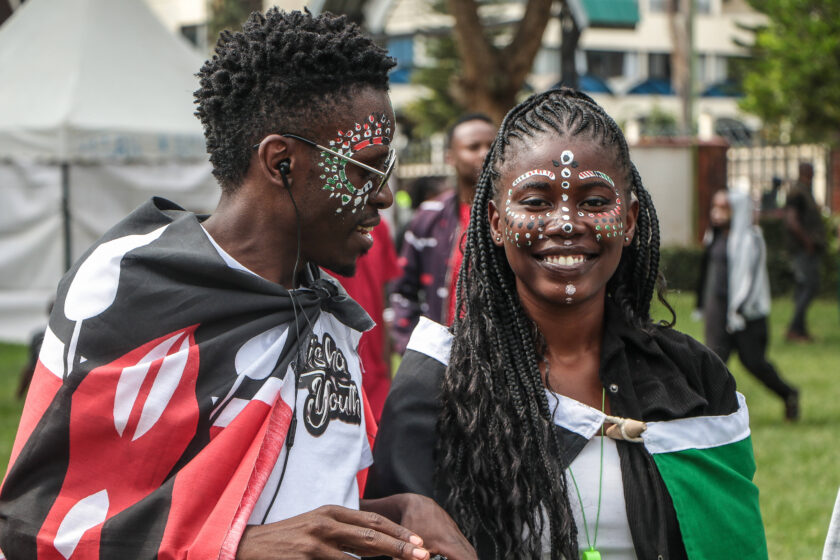 Zwei Mitglieder:innen der Jugendprotestbewegung in Kenia auf einer Gedenkfeier. Sie haben sich kenianische Flaggen um die Schultern gebunden und lächeln.