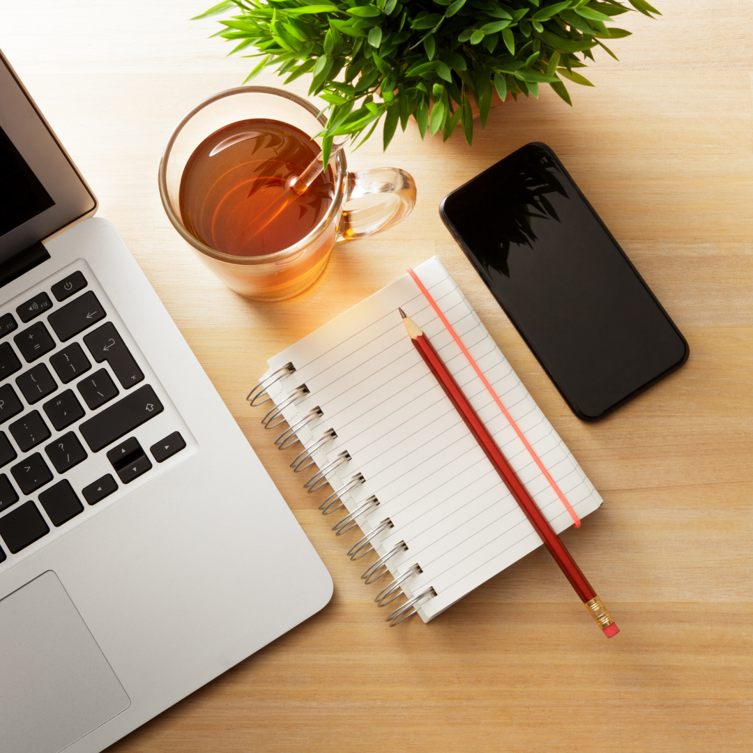 Draufsicht auf einen Schreibtisch. Man sieht die Ecke eines Laptops, einen Notizblock, eine Tasse Tee, ein Smartphone und eine kleine Grünpflanze.