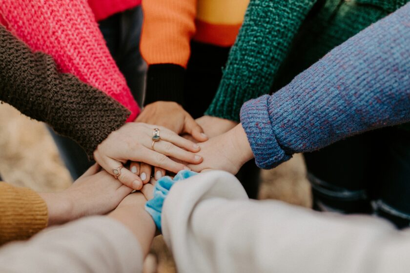 acht Menschen in bunten Pullovern legen ihre Hände übereinander als Zeichen von Zusammenhalt und Solidarität