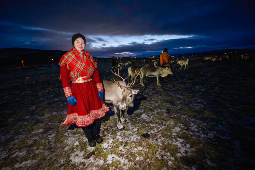 eine junge Sámi-Frau in warmen roten Wollkleidern steht neben ihrer Rentierherde und lächelt in die Kamera