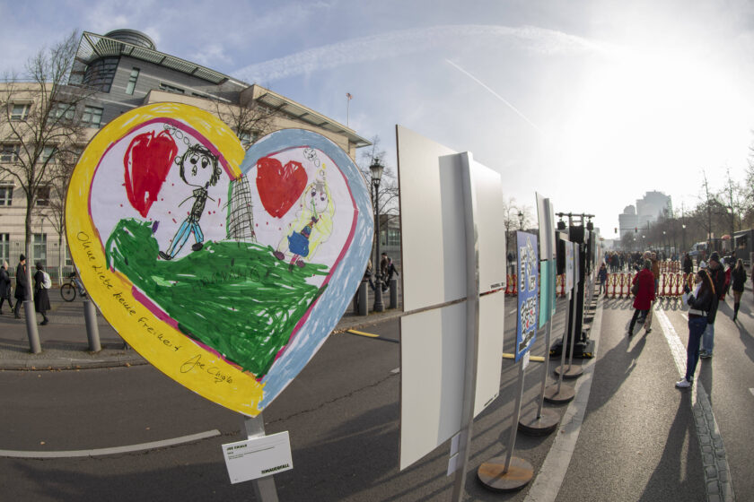 eine Reihe von Plakaten entlang des Verlaufs der Berliner Mauer in Berlin; das Vorderste ist herzförmig und zeigt zwei vermutlich von einem Kind gemalten Menschen mit Herzen. Auf dem Plakat steht: Ohne Liebe keine Freiheit.