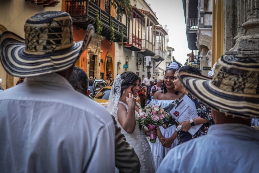 Eine Hochzeit in Kolumbien: Die lächelnde Braut spricht mit zwei Frauen, Männer mit traditionellen Hüten stehen - der Kamera den Rücken zugewandt - daneben. Im Hintergrund sieht man farbenfrohe, mehrstöckige Wohnhäuser.