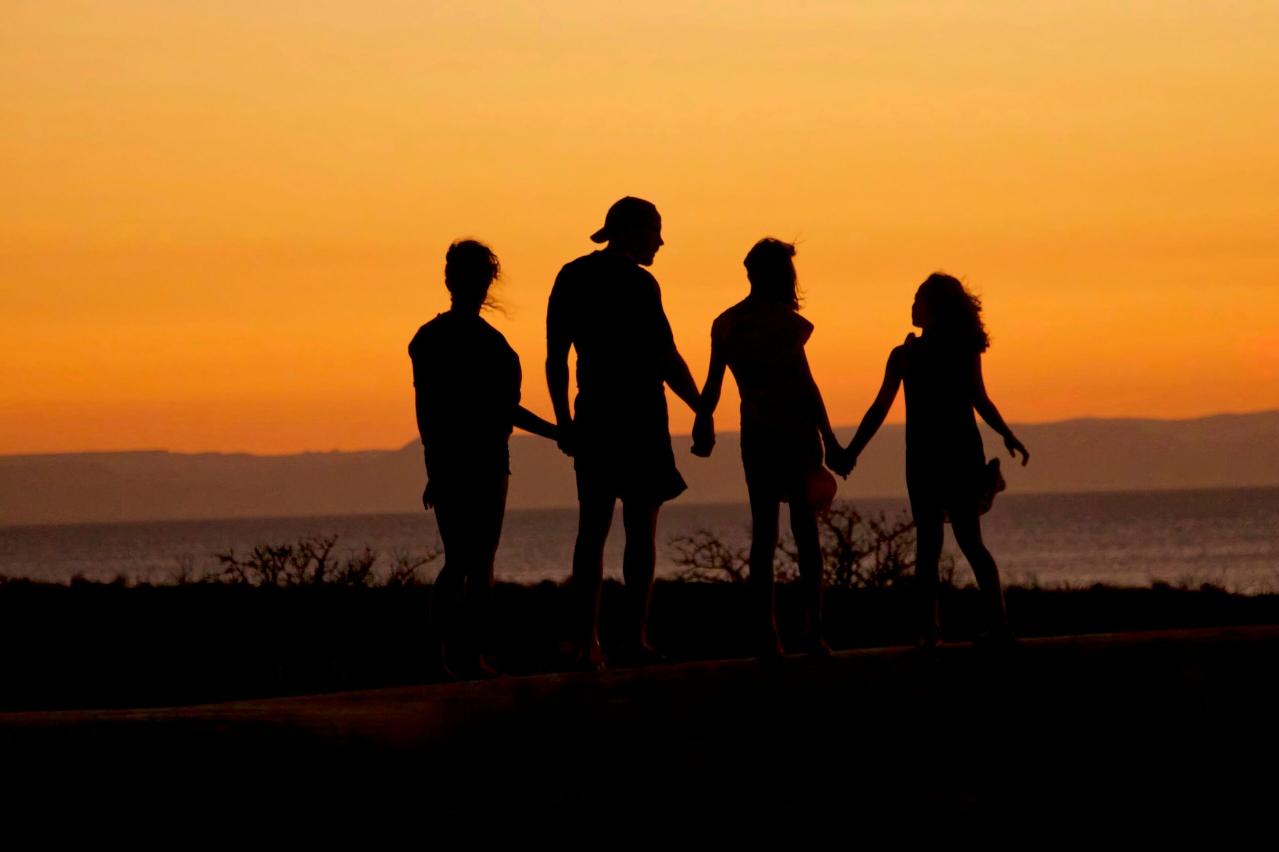 die Silhouette von vier Personen, die sich an den Händen halten, gegen einen orangefarbenen Sonnenuntergang