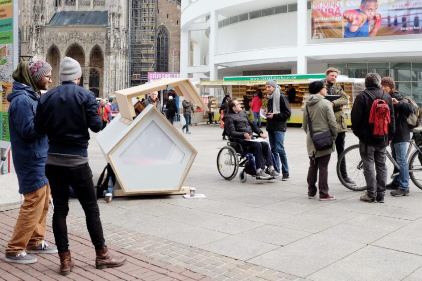 Menschen begutachten ein Ulmer Nest in der Ulmer Innenstadt. Das Nest ist ein pentagonales Konstrukt aus Holz.