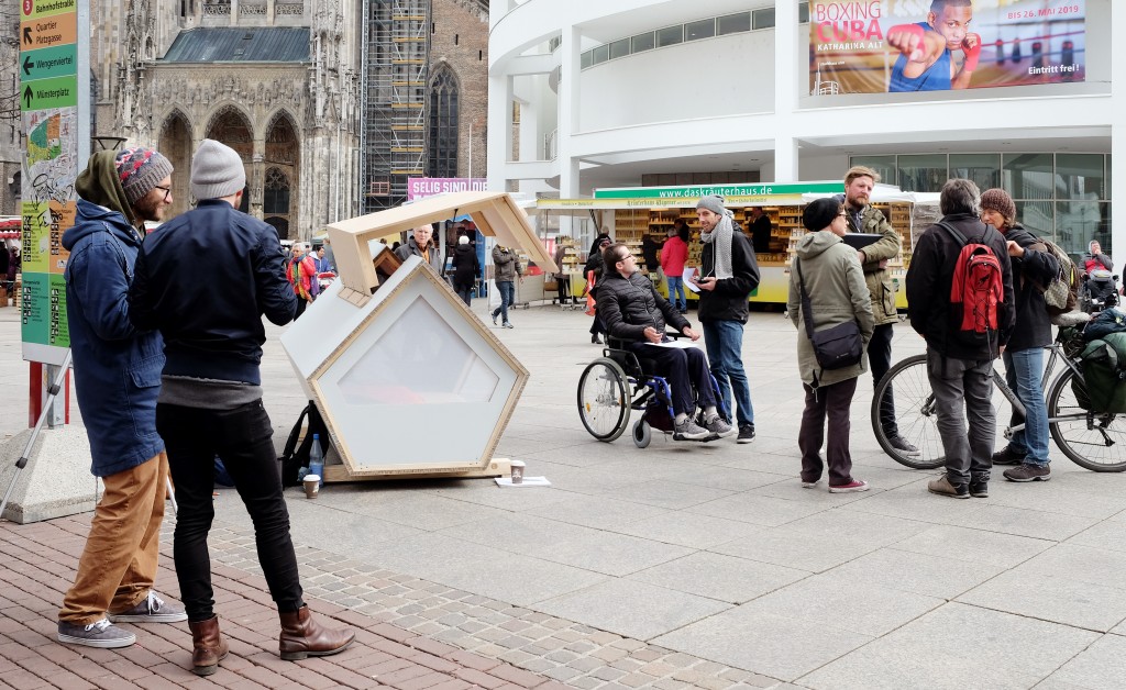 Menschen begutachten ein Ulmer Nest in der Ulmer Innenstadt. Das Nest ist ein pentagonales Konstrukt aus Holz.