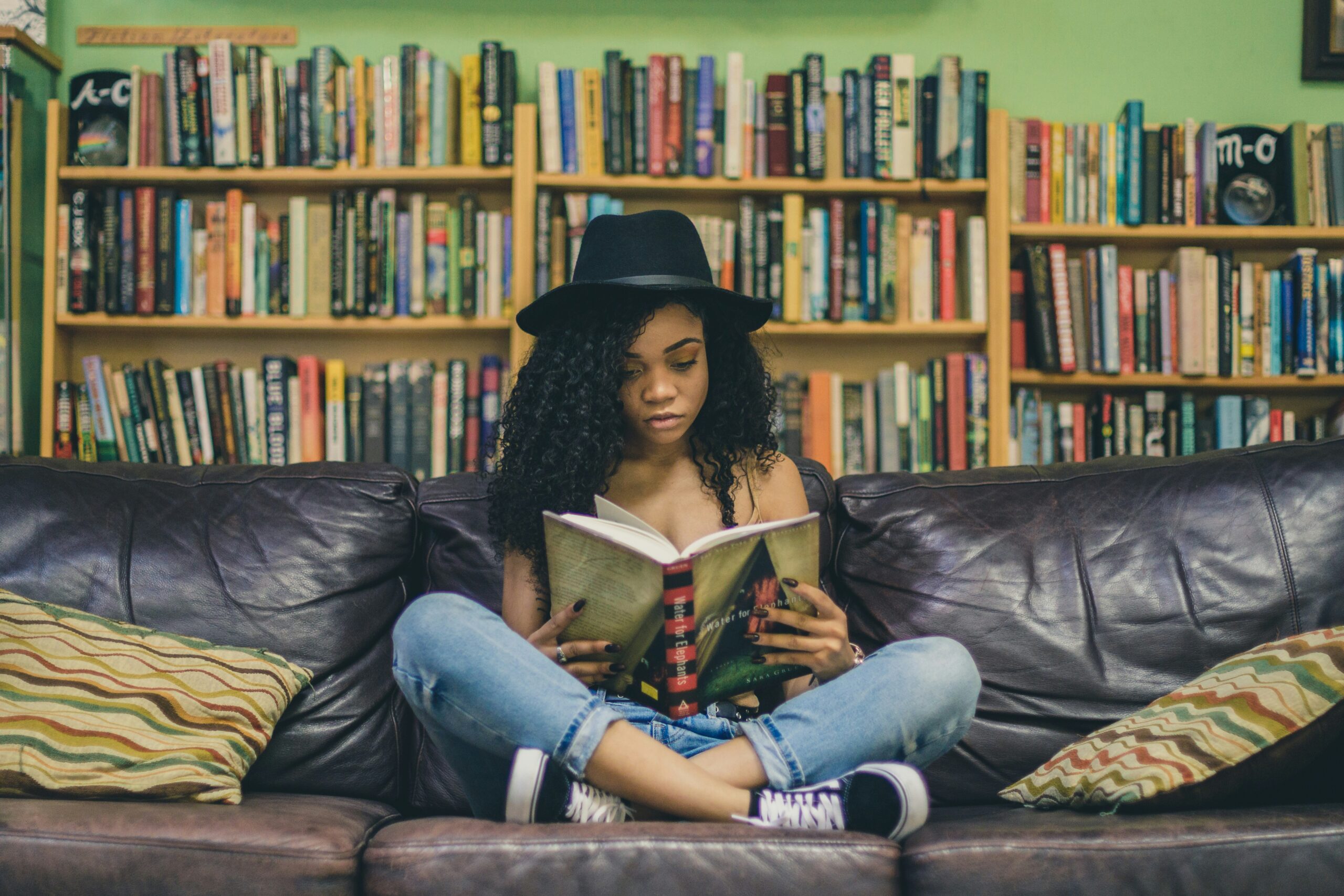 eine Schwarze junge Frau sitzt im Schneidersitz auf einem Sofa und liest ein Buch. Im Hintergrund stehen Regale voller Bücher.