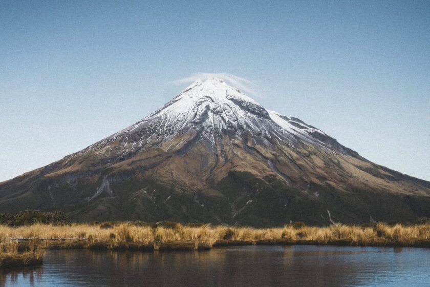 Ein schneebedeckter Berg spiegelt sich in einem stillen See unter einem klaren blauen Himmel, Mount Taranaki,