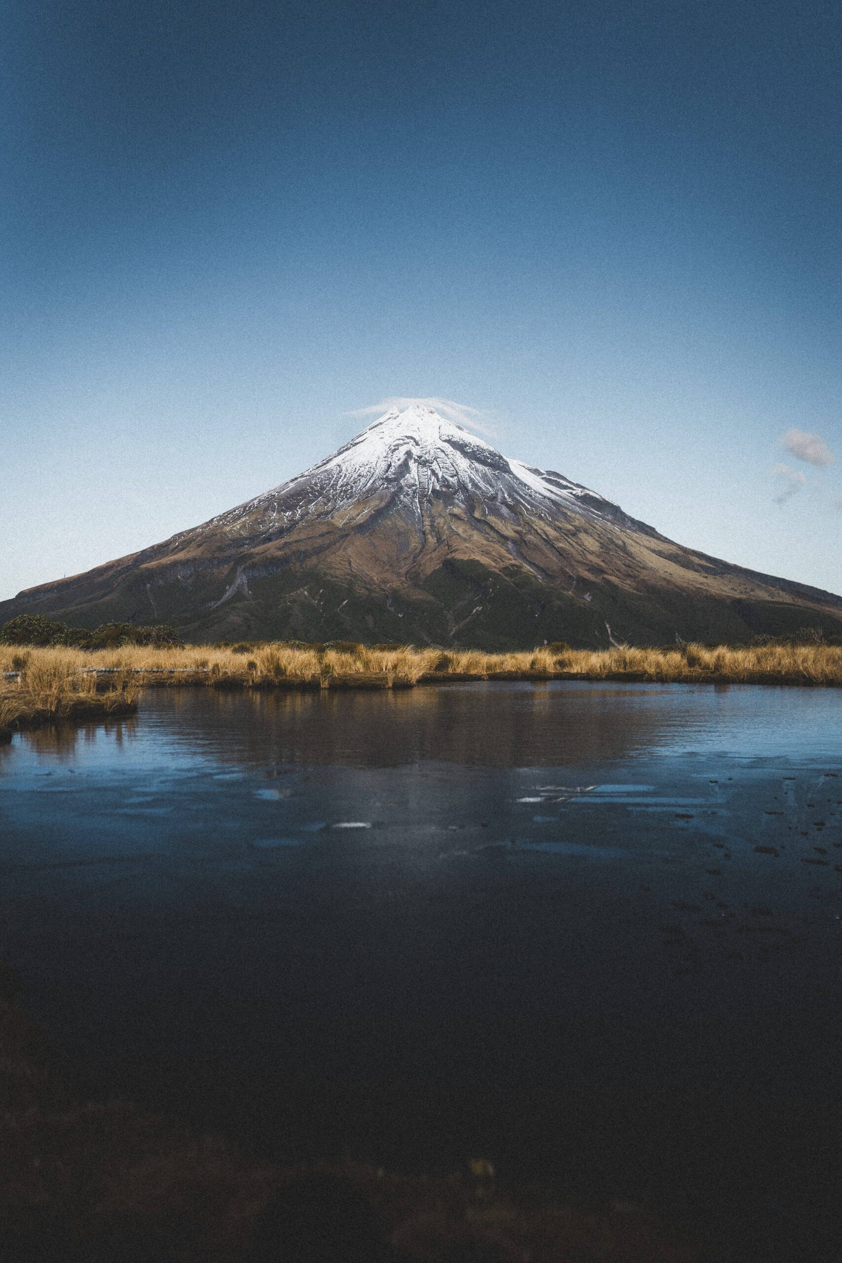 Ein schneebedeckter Berg spiegelt sich in einem stillen See unter einem klaren blauen Himmel, Mount Taranaki,