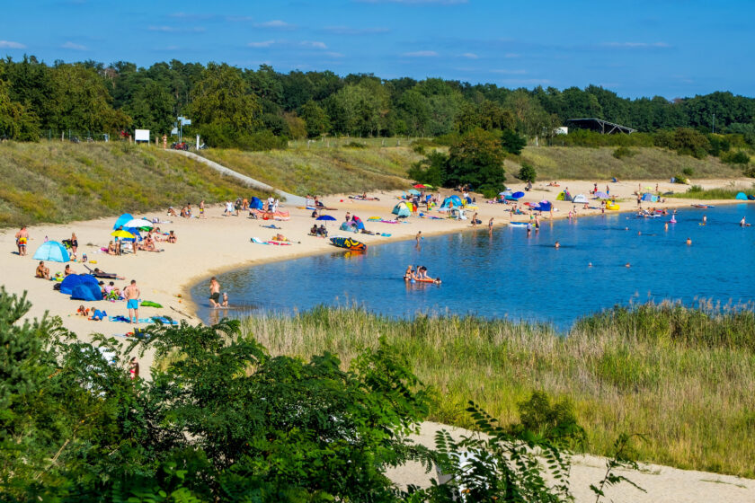 Das Bild zeigt einen Badesee mit Strand und Wiese, an dem viele Leute liegen und schwimmen gehen
